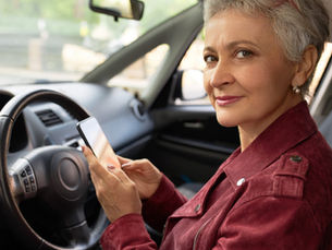 Um senhor ou senhora (acima de 60 anos) sorridente, sentado(a) ao volante de um carro moderno, em um ambiente claro e ensolarado.